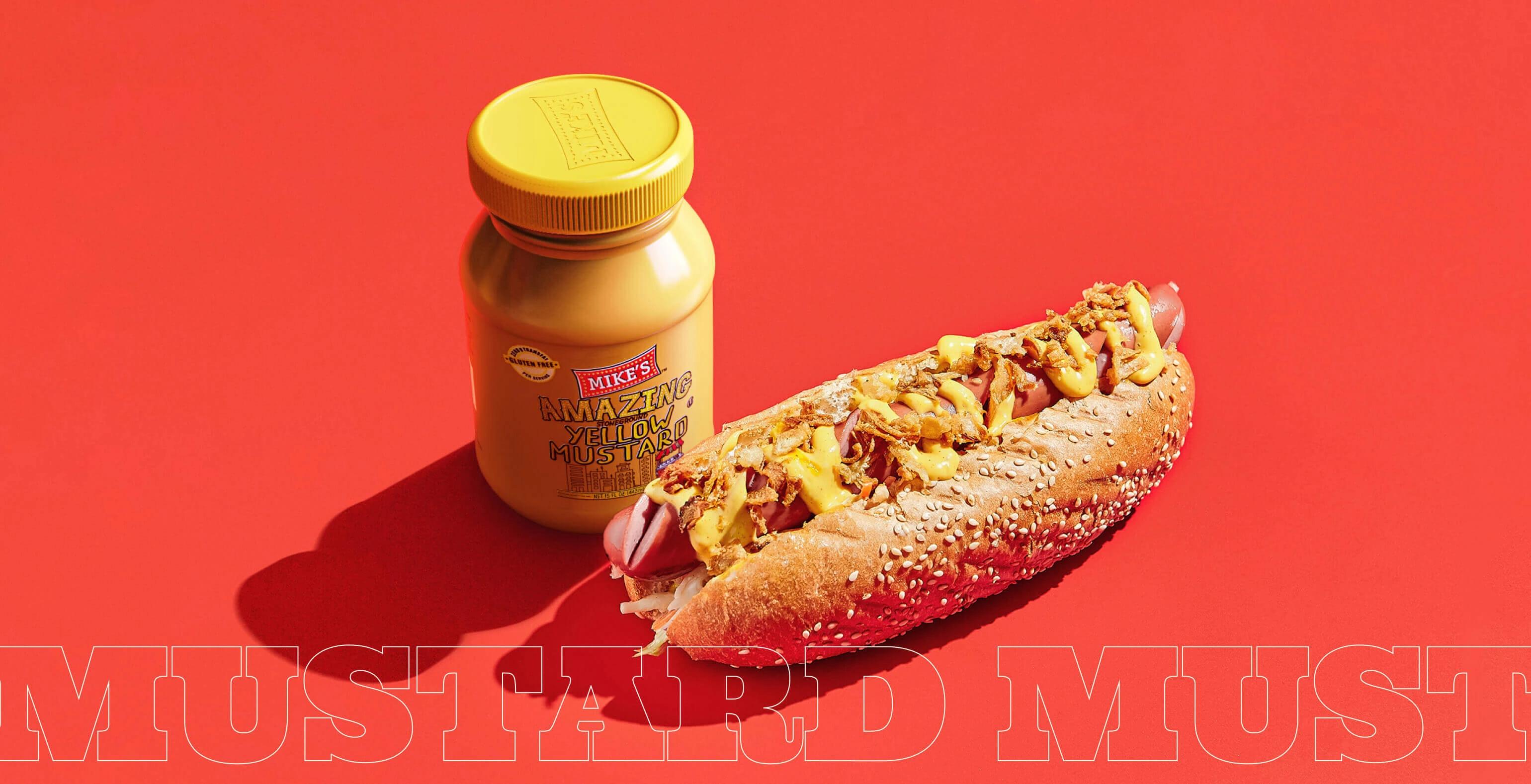 A bottle of Mike's Amazing Mustard next to a tasty sandwich.